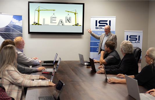 A group of ISI Solar employees meet around a large wooden table. They are all looking toward the back of the table where a man points to a large screen on the wall. "LEAN" is spelled out on the screen and it looks like he is doing a training on best practices.