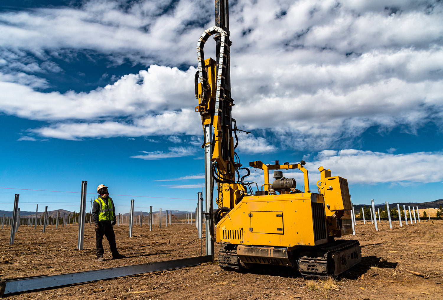This photo captures the beginning stages of a solar panel site.  On the right, a large yellow machine is helping set the supports for the solar panels.  A man wearing a safety vest and hard hat stands to the left watching the machine set the post.