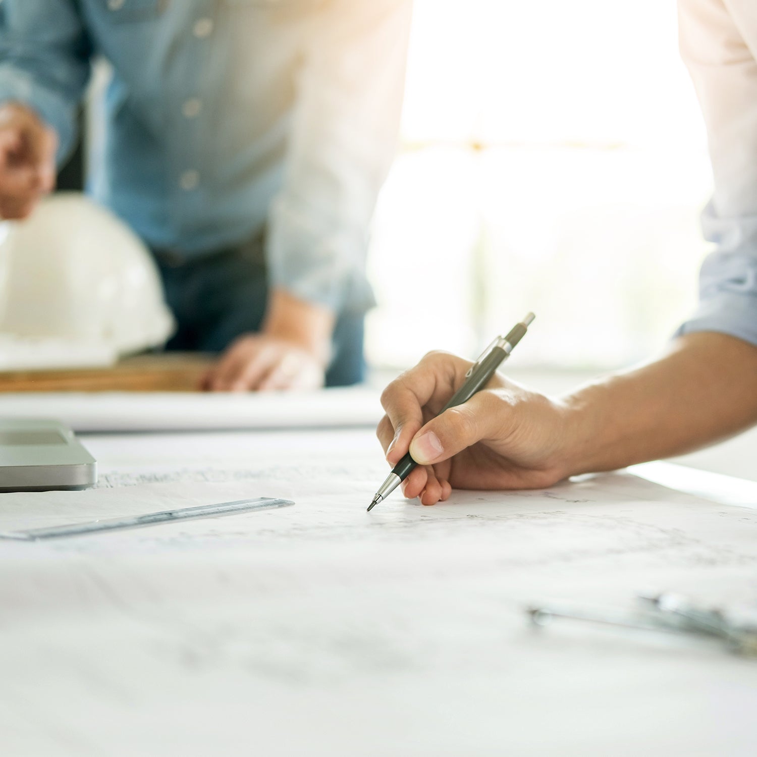 Person holding a pen and working on blueprints with colleagues in the background.