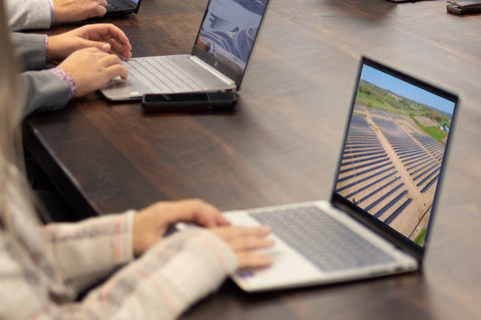 A close up photo of 3 people on their laptops.  Their laptops are on a brown wooden table.  The woman's laptop is the closet to the front of the photo and she rests her hands on her laptop.  A photo of a solar field is on her screen.