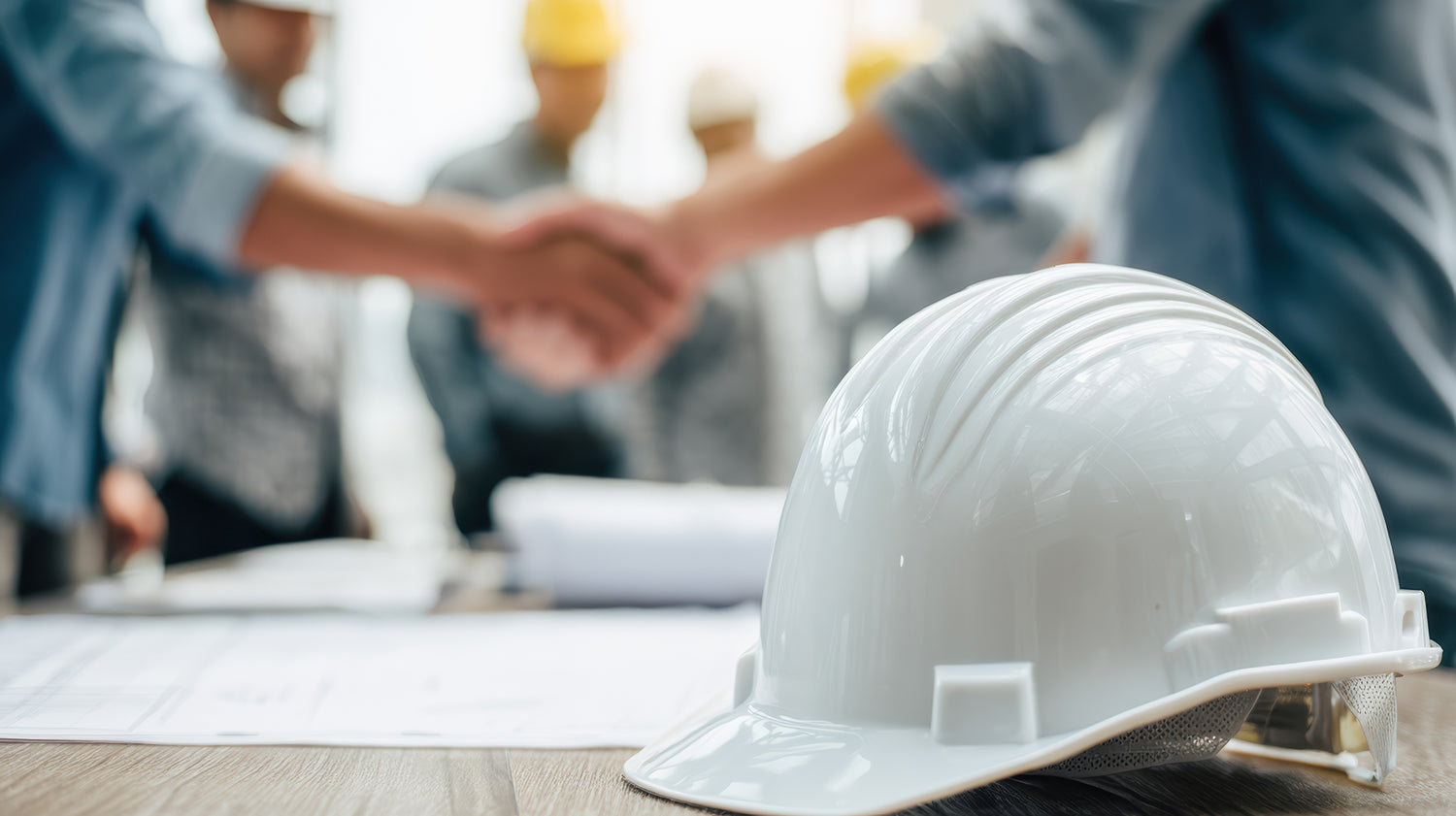 Two people shaking hands with a white hard hat in the foreground on a blurred background of people and documents.