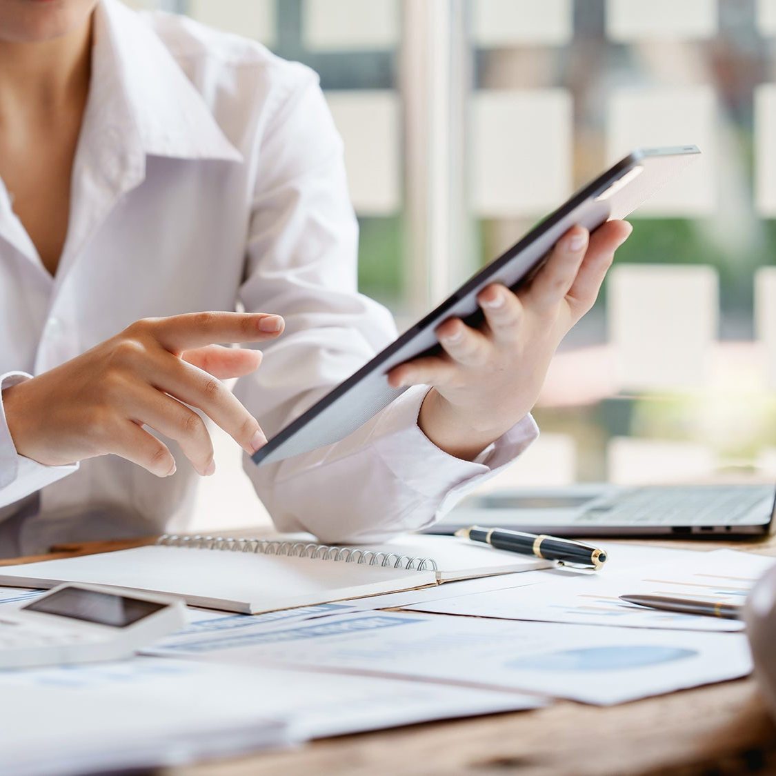 Person using a tablet at a desk with papers and a laptop in the background