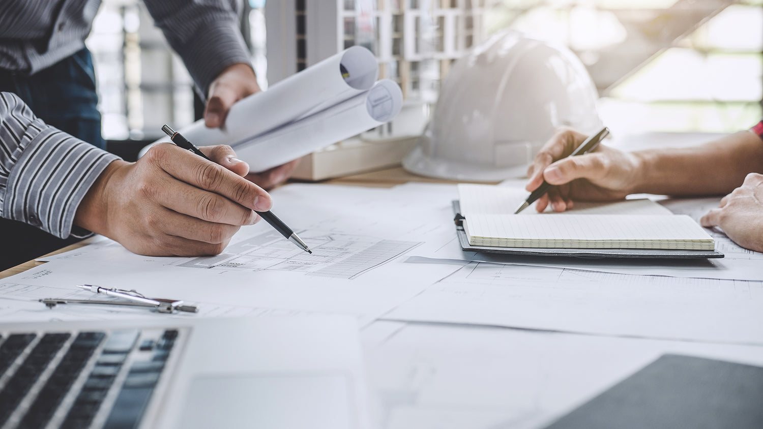 Two people working on blueprints and documents at a desk with a laptop.