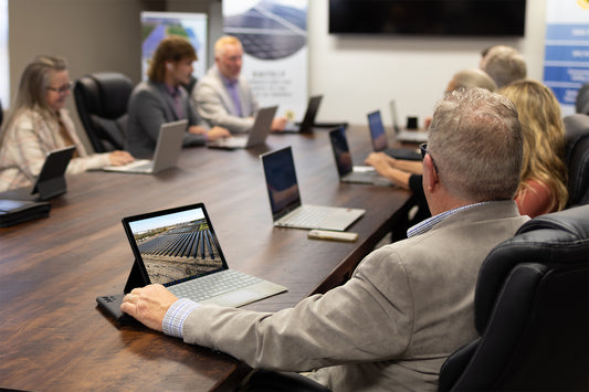 A group of ISI Solar employees meet at a brown wooden table.  In focus, we see the back of a man in a grey suit.  A solar field is on his screen.  Everyone else is out of focus, but they all have their laptops in front of them.