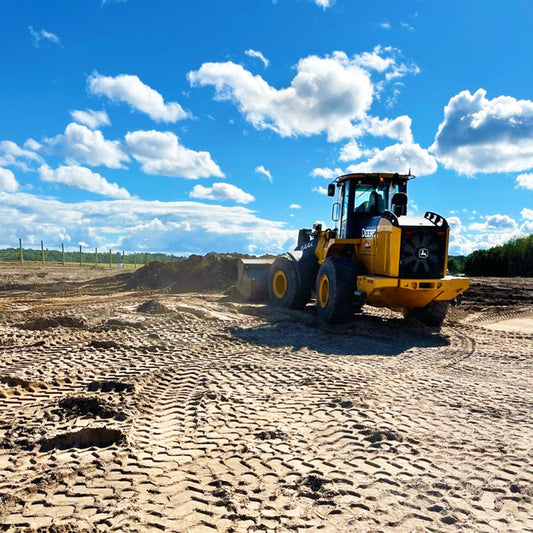 A bright blue sky with white puffy clouds hovers over brown dirt covered in machinery tire prints.  A yellow tractor takes up the right side of this photo as it pushes dirt to the back.