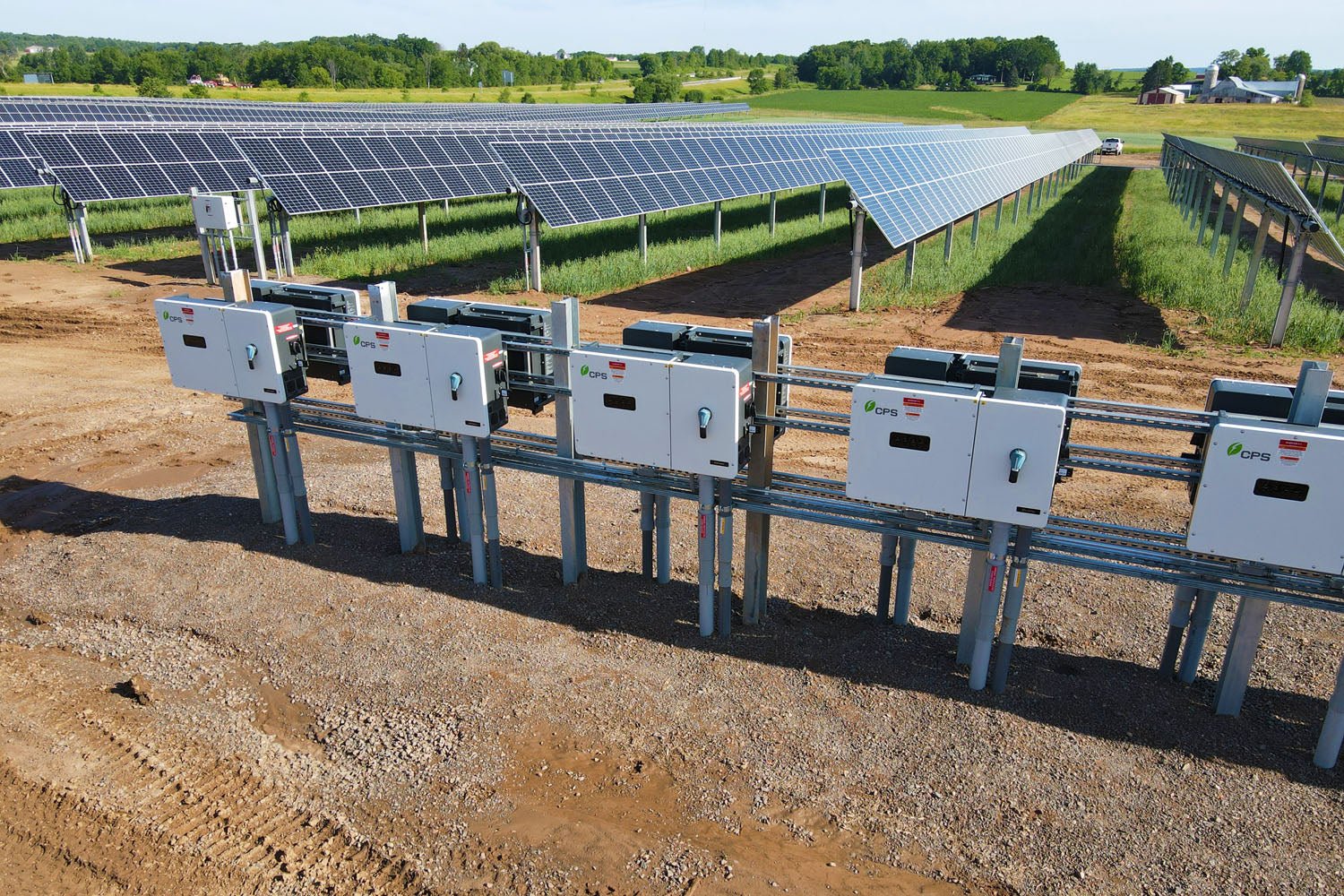 A solar panel site with the control system on gravel in the front of the photo.  Behind the controls, the solar panels are neatly in rows in a grassy field.  There is a small farm place in the background.