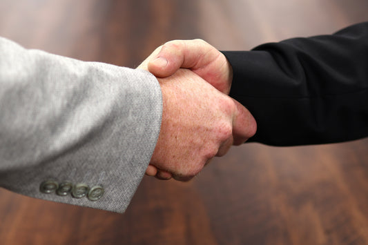 A close up photo of two men shaking hands.  The man on the left wears a gray suit and the man on the right wears a black suit.  You can only see their hands and first few inches of their arm.