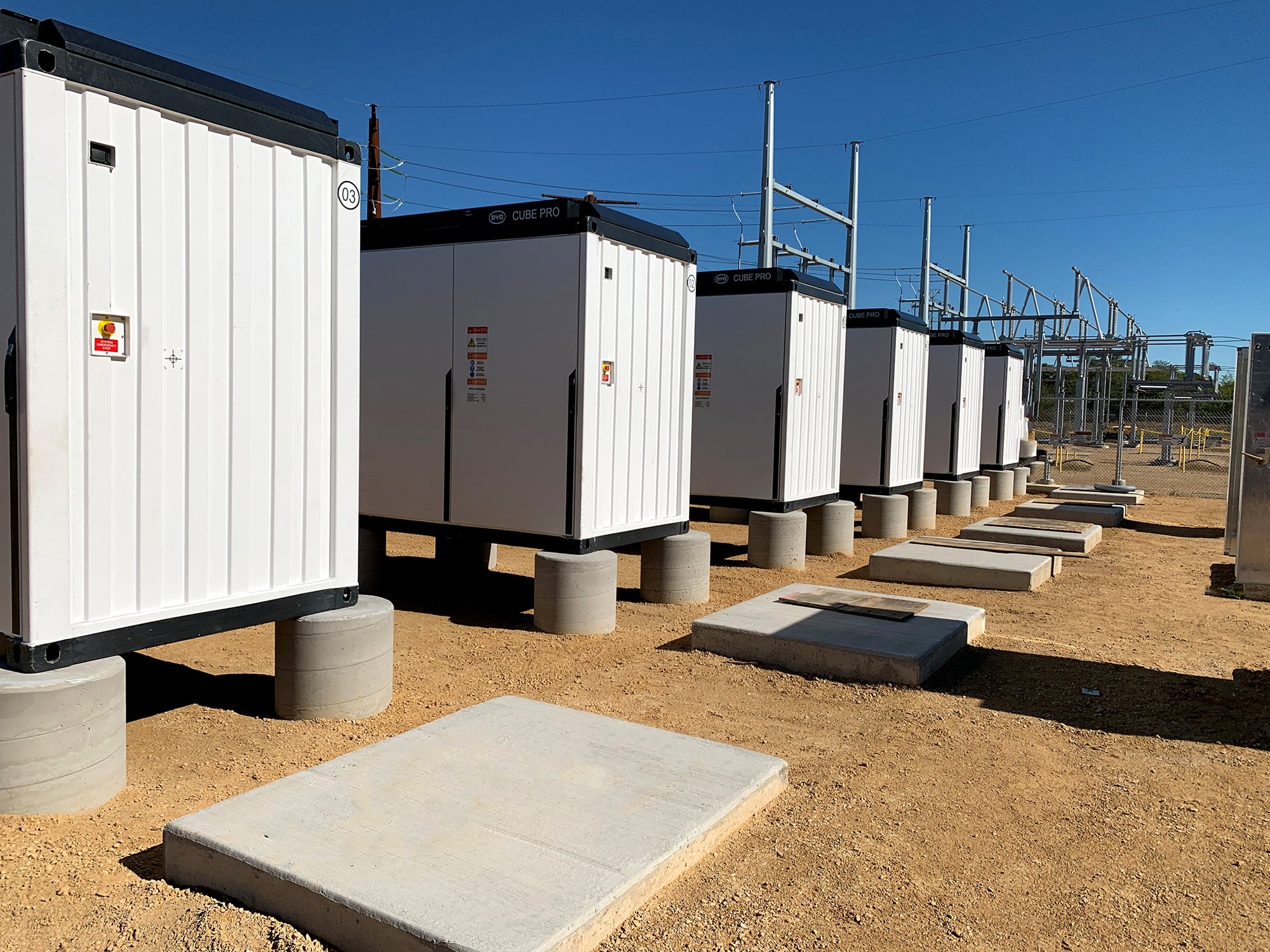 Row of large battery storage units in an outdoor setting with a clear blue sky.