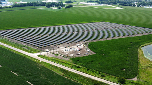 This solar site is surrounded by bright green grass.  There dirt parking lot next to the solar panels.  It looks like a few portable buildings and several  trucks are in the parking lot.