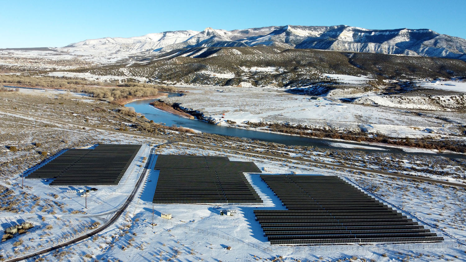 A solar site at the base of the mountains.  A light snow semi covers the ground and mountains in the background.  A small river separates the solar panels and the mountains.