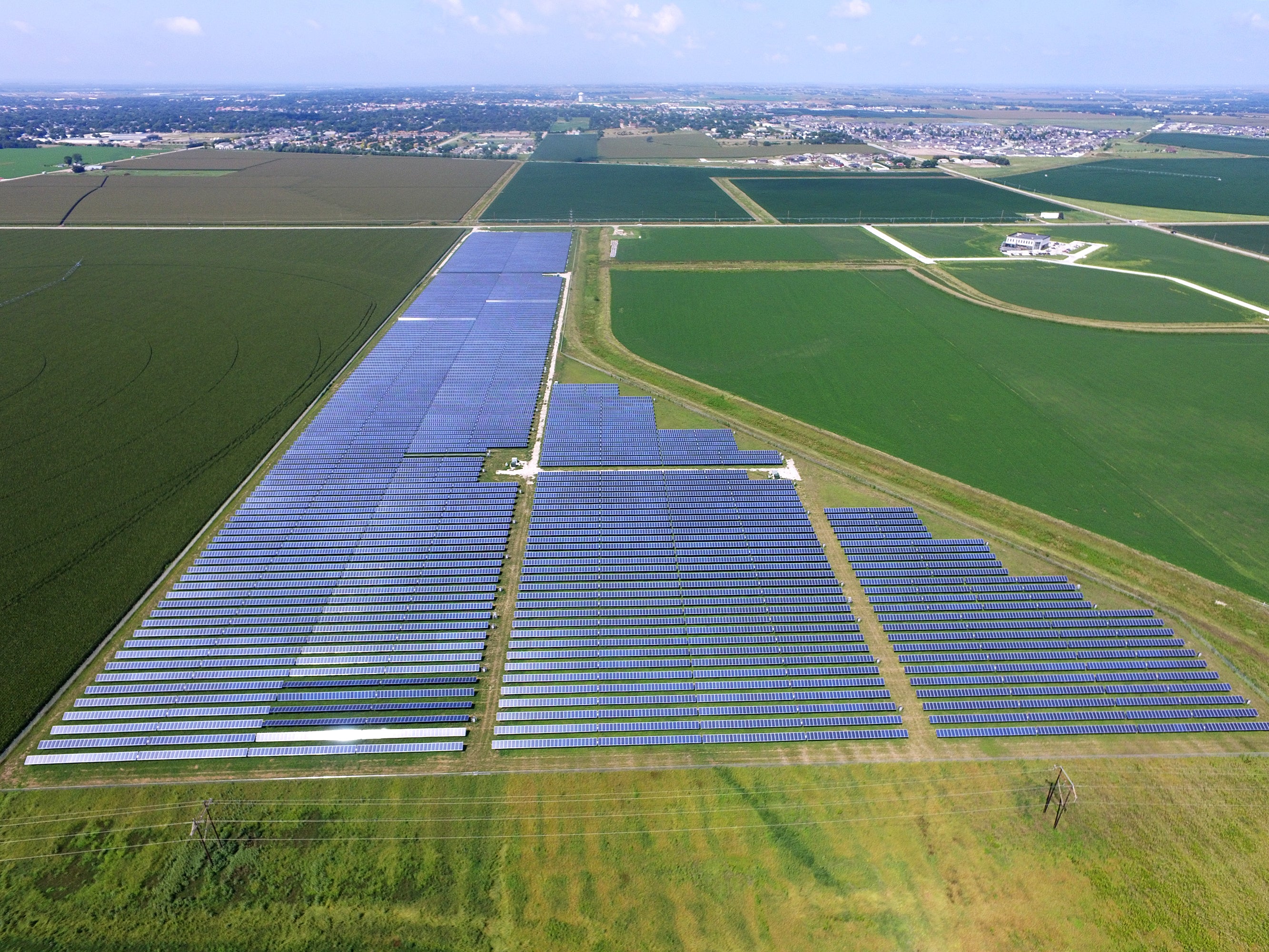 This solar site has 3 rows.  The one on the left is the longest.  The middle is a medium length.  The right is the smallest row.  This site is in the middle of green fields.  In the distant background, you can see a city.