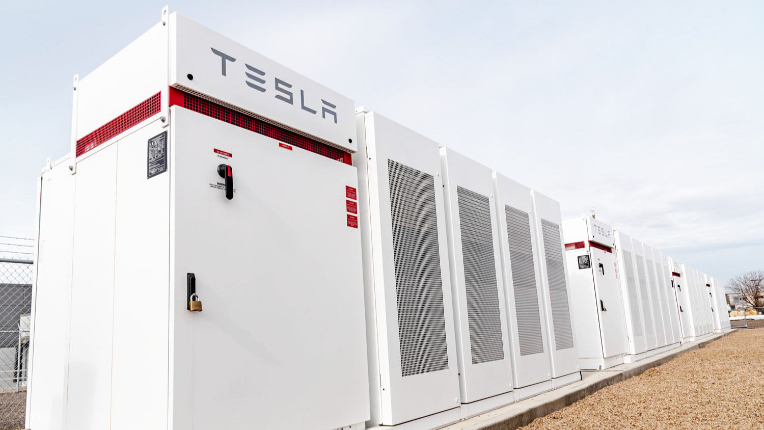A close up of white BESS.  They are set on concrete and there is a thick layer of gravel below.  They sky above is a grayish white - almost the same color as the BESS units. The Tesla logo is on the first unit you see in the top left.  