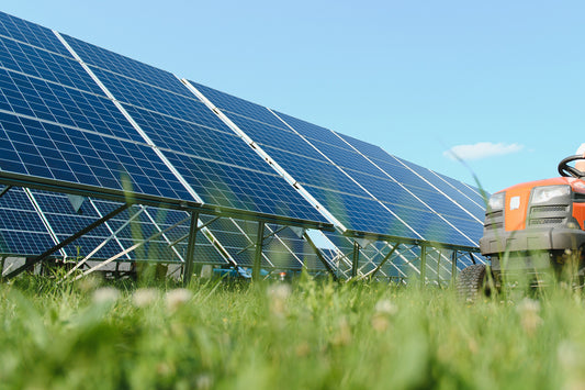 The photo is taken from the ground.  You see blurry grass with solar panels in focus in the background.  And orange and black lawnmower is starting to come into the frame from the right side of the photo.