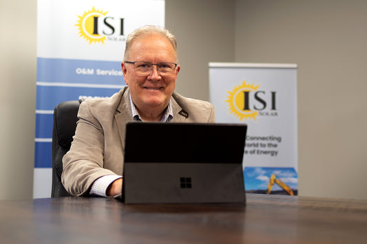 A man sits at a wooden table and looks into the camera.  He has a tablet in front of him.  He wears glasses and is smiling.  ISI Solar banners are out of focus in the background behind him.